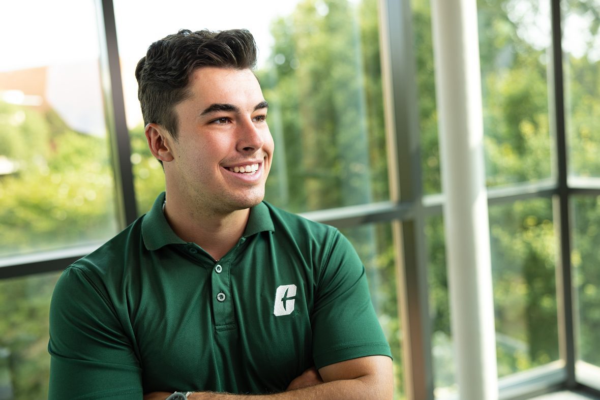 A smiling student in a green UNC Charlotte polo shirt stands indoors near large windows with trees visible outside.
