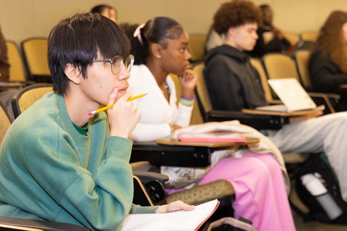 Students sit in tiered lecture hall seats, taking notes and listening attentively, with notebooks, pencils, and laptops in front of them.