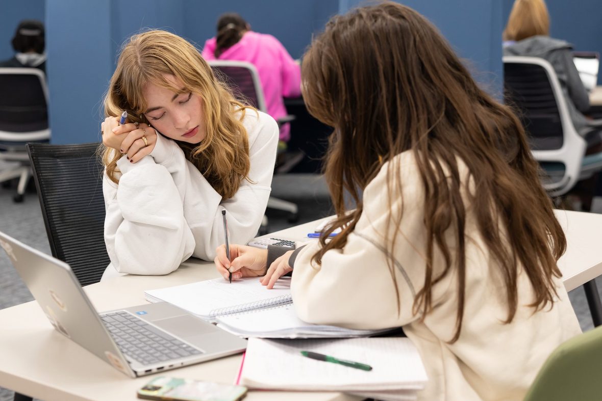 Two students in a study area sit at a small table with open notebooks and a laptop, one writing while the other leans on her arm watching.